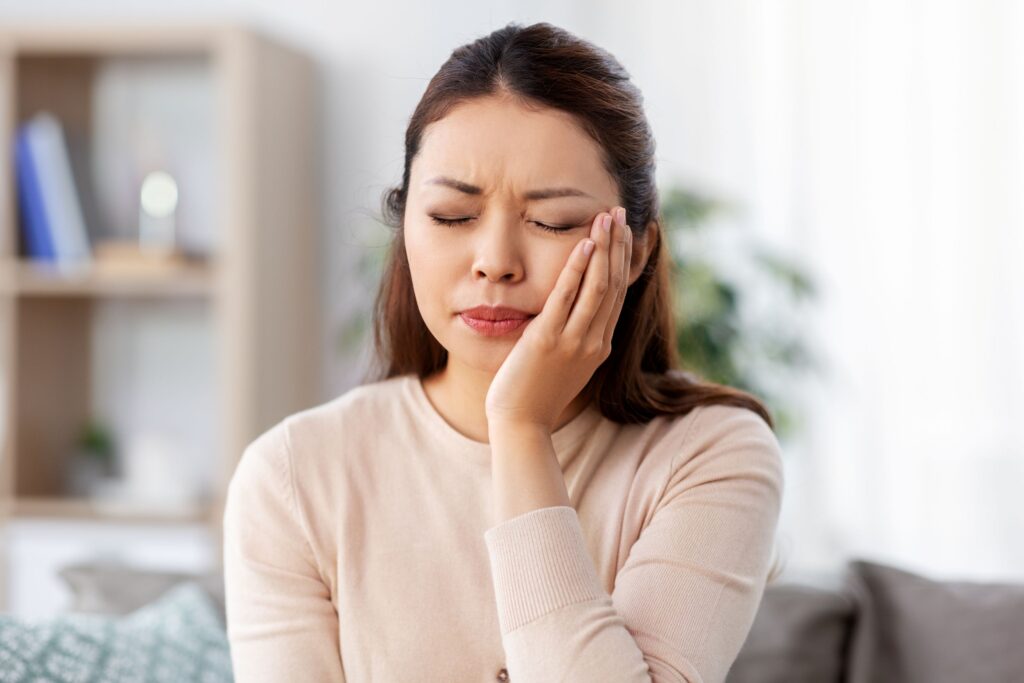 Woman in beige sweater with eyes closed holding hand to face in pain